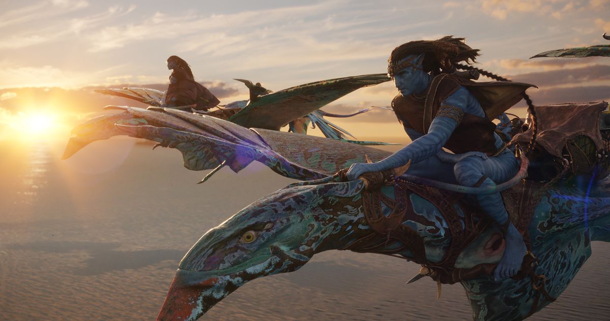 Na’vi mates Neytiri (Zoe Saldaña) and Jake (Sam Worthington) fly on their mottled blue banshee mounts above the clouds with a sunset behind them in Avatar: The Way of Water