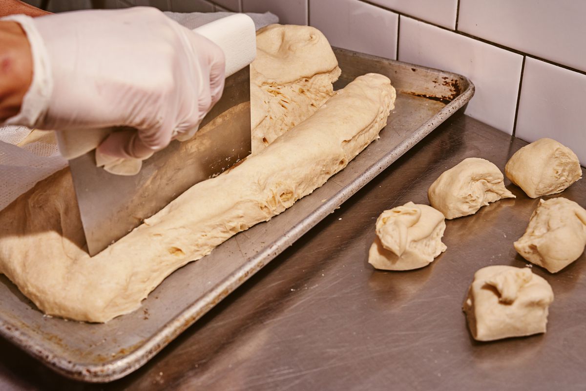 A gloved hand divides a smooth, beige-colored dough into small pieces using a metal bench scraper
