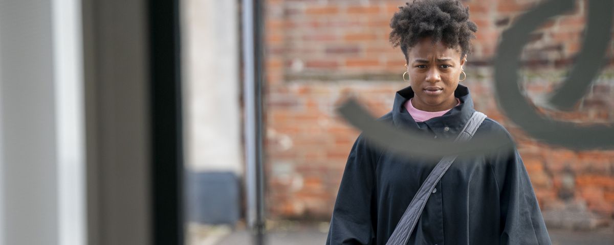 A young Black woman (Bukky Bakray) in a black coat and bright red gloves stands outside of a window looking in in a scene from Netflix’s The Strays