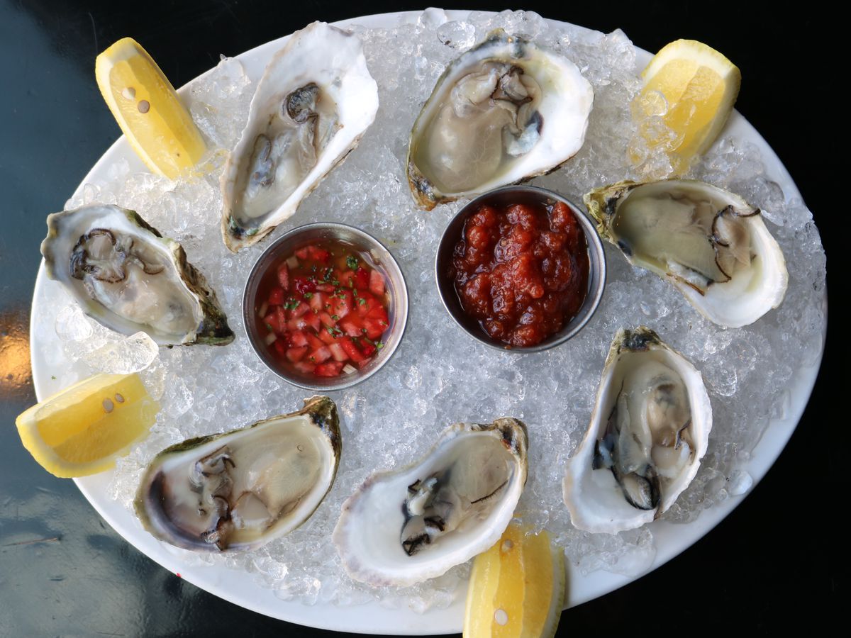An ice-filled tray of oysters interspersed with lemon wedges and two cups filled with a red garnishing sauce.