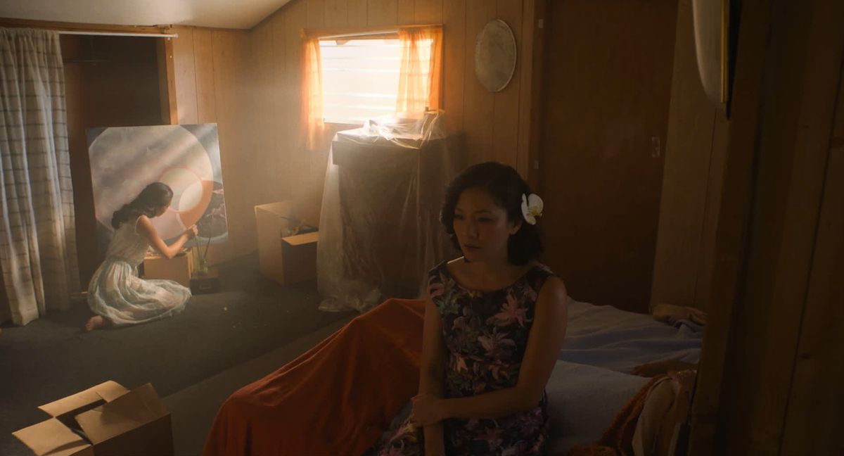 In “I Was A Simple Man,” Constance Wu sits on a bed in the foreground while sunlight peers through a window onto another woman painting in the background of the same room.