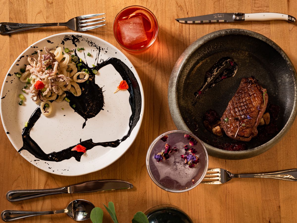 A white plate with calamari, black sauce, and bright orange nasturtiums; a black plate with seared duck breast; a purple cocktail; a red cocktail with a large square ice cube.