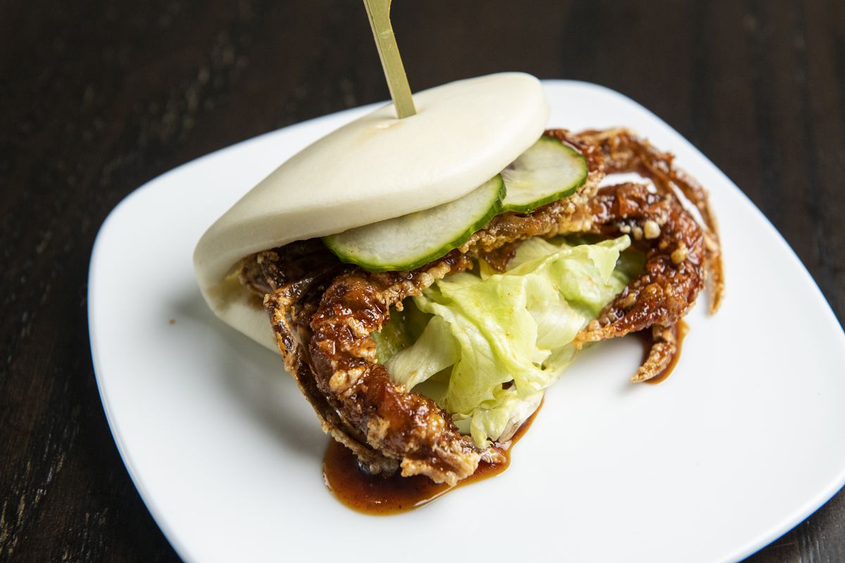 Battered soft shell crab legs stick out the sides of a soft bao bun. It’s garnished with sliced cucumbers and lettuce.