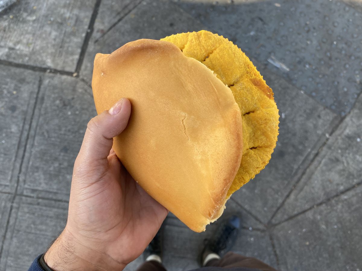 A hand clutches a yellow patty tucked into a piece of coco bread.