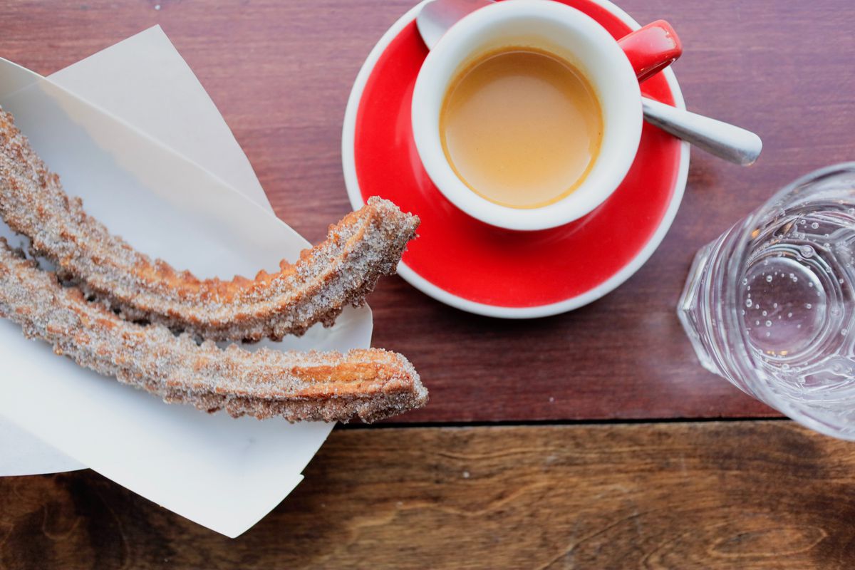 Two churros on a white tray next to a coffee in a mug on top of a red saucer next to a glass of water, all on a wooden table
