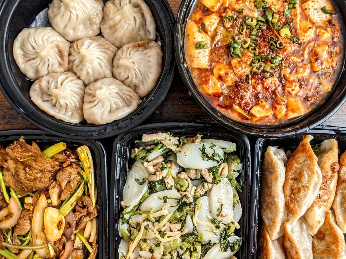 Overhead view of two round and three rectangular black plastic takeout containers of Chinese food, including two kinds of dumplings, ma po tofu, oval rice cakes with greens and slivers of pork, and more.