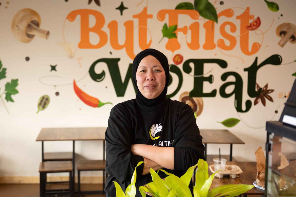 A woman wearing a head covering stands with her arms crossed in front of a sign that reads, “first, we eat.”
