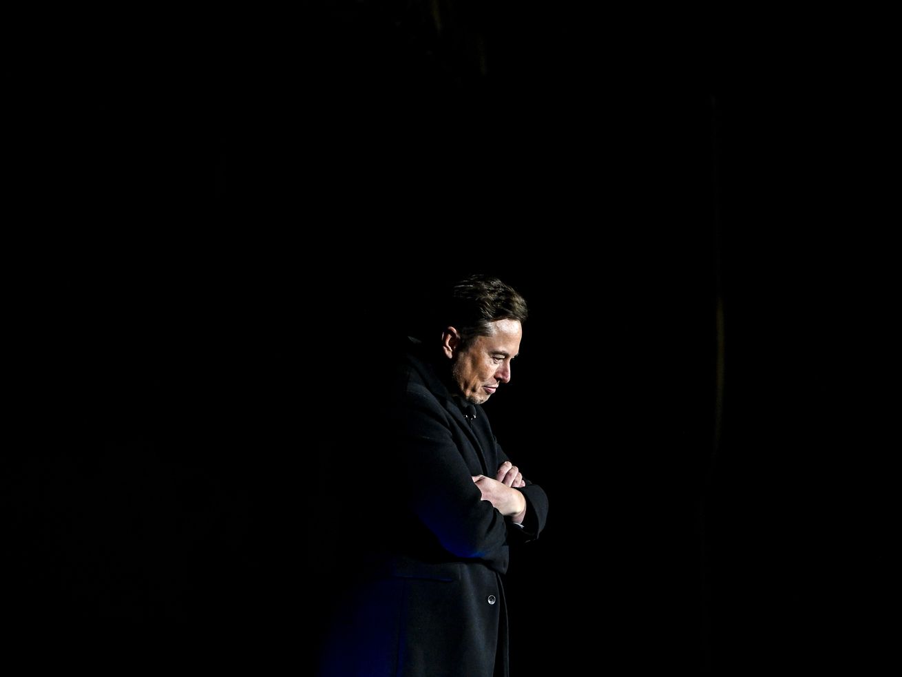 Elon Musk standing lit on a dark stage with his arms folded.