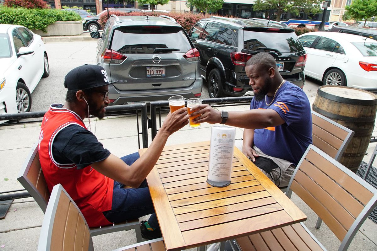 Two Black men sitting at an outdoor patio table clink their beer glasses.