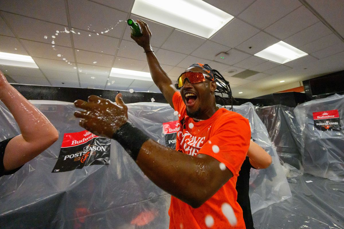 Orioles player Jorge Mateo, wearing an orange “Take October” shirt and goggles over his eyes, shouts as he flings beer from a bottle around the plastic-covered clubhouse