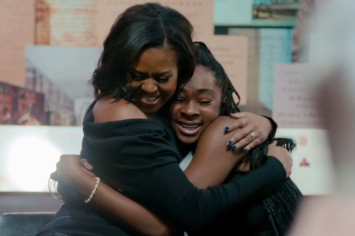 Image from the Netflix documentary, 'Becoming' (2020). Michelle Obama hugs a young black girl tightly. The girl appears to be crying whilst Michelle smiles.