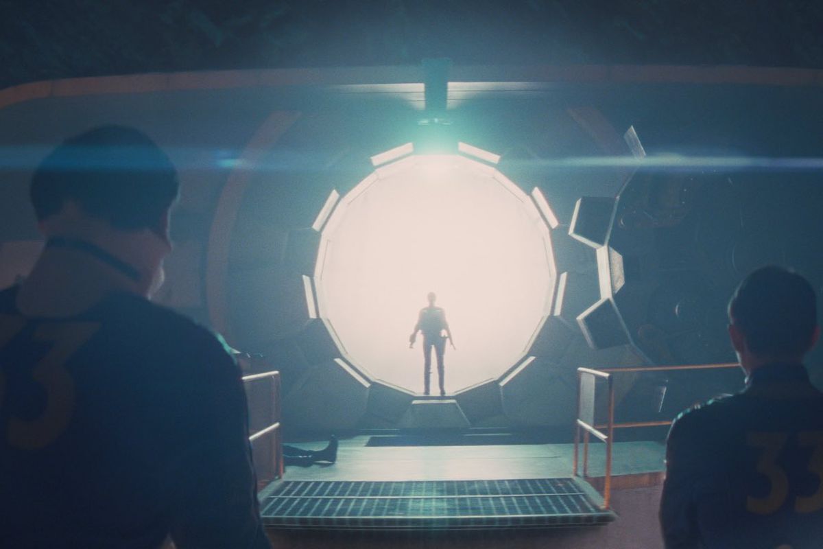 in a scene from the Fallout TV series, a silhouette stands in front of the blinding white light flooding into an underground shelter from the open vault door of Vault 33 while three people in denim jumpsuits with 33 on the back look on.