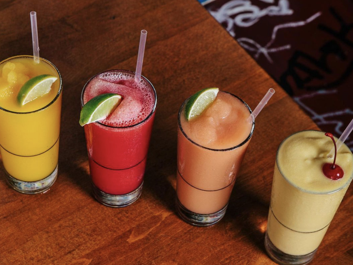 Four frozen drinks in tall pint glasses in a variety of flavors on a wood table.