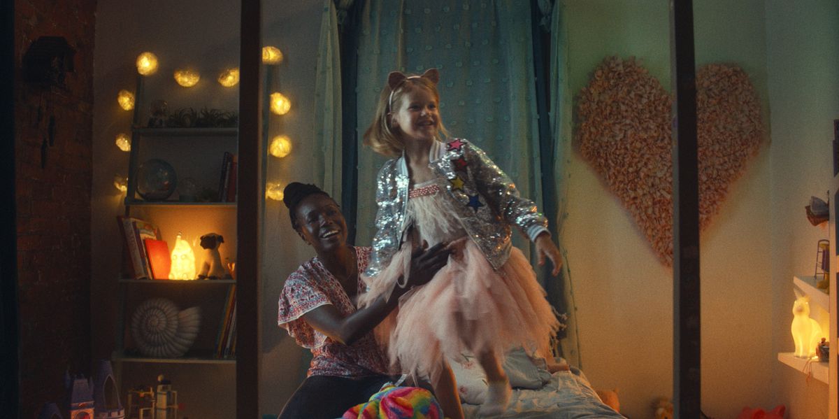 Aisha (Anna Diop), a dark-skinned woman in a colorful pink patterned top, holds the waist of Rose (Rose Decker), a young blonde Caucasian girl wearing a cat-ear headband, silver jacket and pink tutu, as she jumps on a bed in nanny