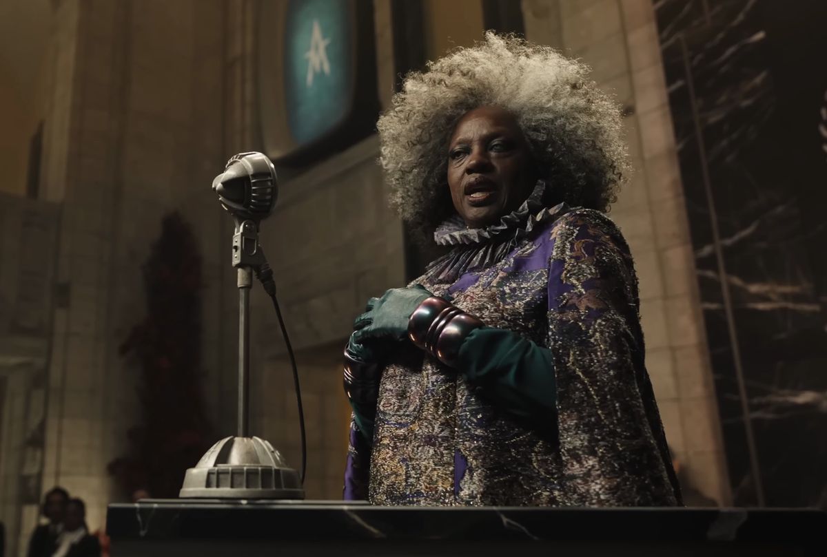 Viola Davis as Dr. Gaul with big frizzy gray hair and a purple gown speaking at a microphone in The Hunger Games: The Ballad of Songbirds and Snakes