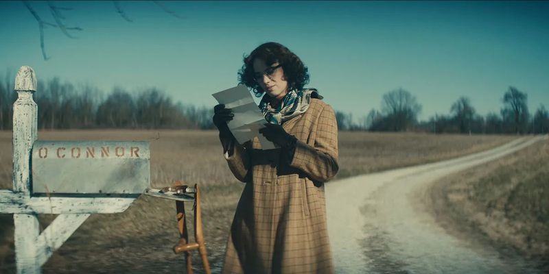 A woman in a long coat and a curly haircut stands next to a mailbox on a desolate road, reading a letter.