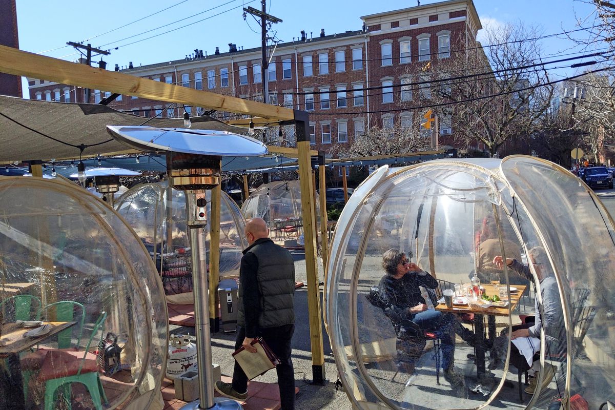 A parking lot with plastic bubbles, each with one table, and brownstones in the background.