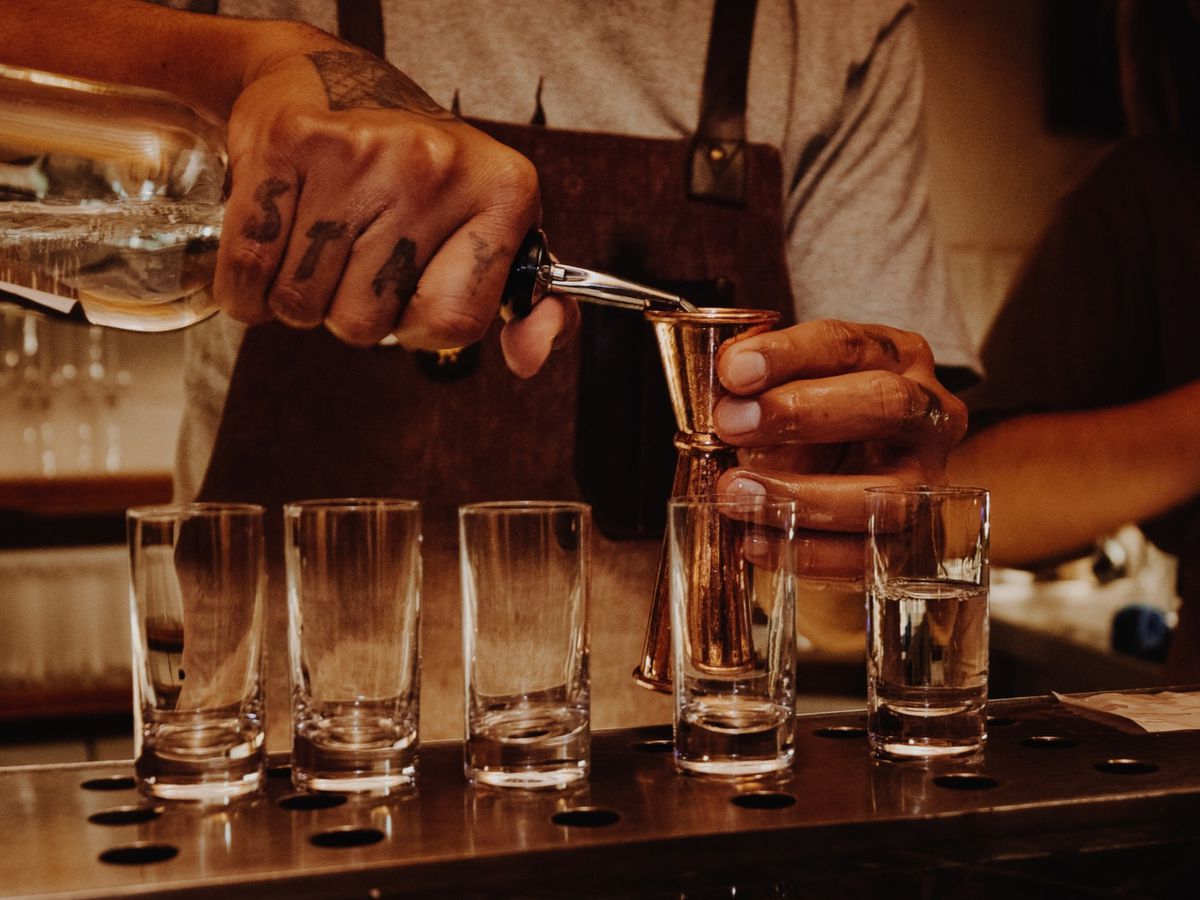 A bartender pours a bottle into a jigger, behind glasses lined up for drinks