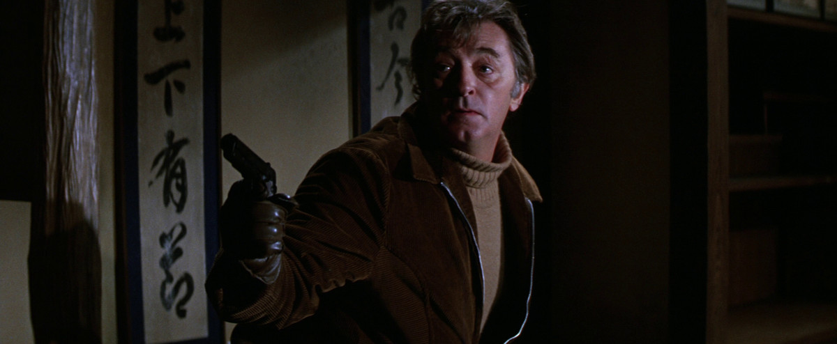 Robert Mitchum, wearing a turtleneck and a jacket, holds a gun next to Japanese script in The Yakuza.