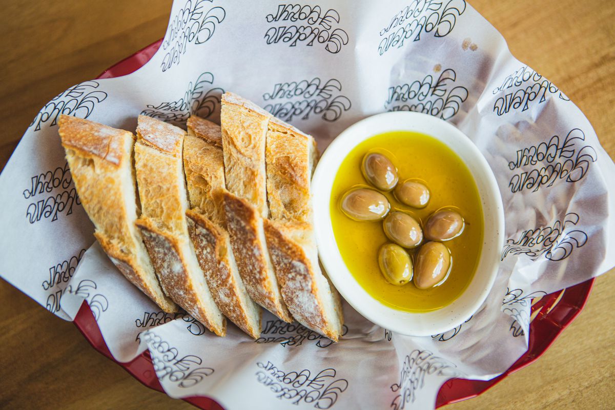A wax-paper-lined tray of sliced bread and small bowl of olive oil and olives.