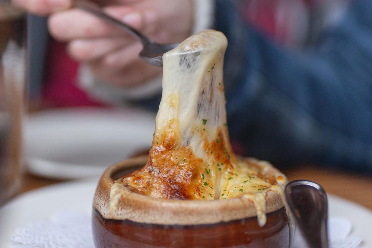 A hand holds up a spoon full of cheese, stretching down to a clay pot of onion soup.