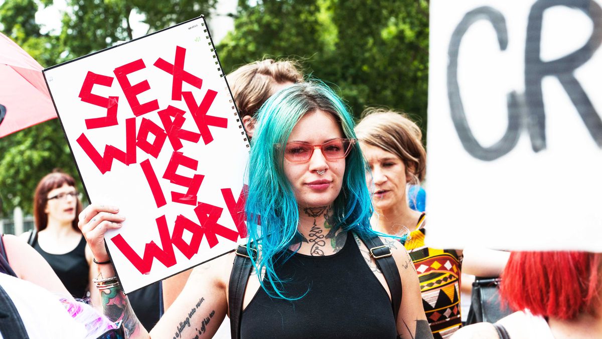 A woman with blue hair holds a sign that says “Sex Work Is Work.”