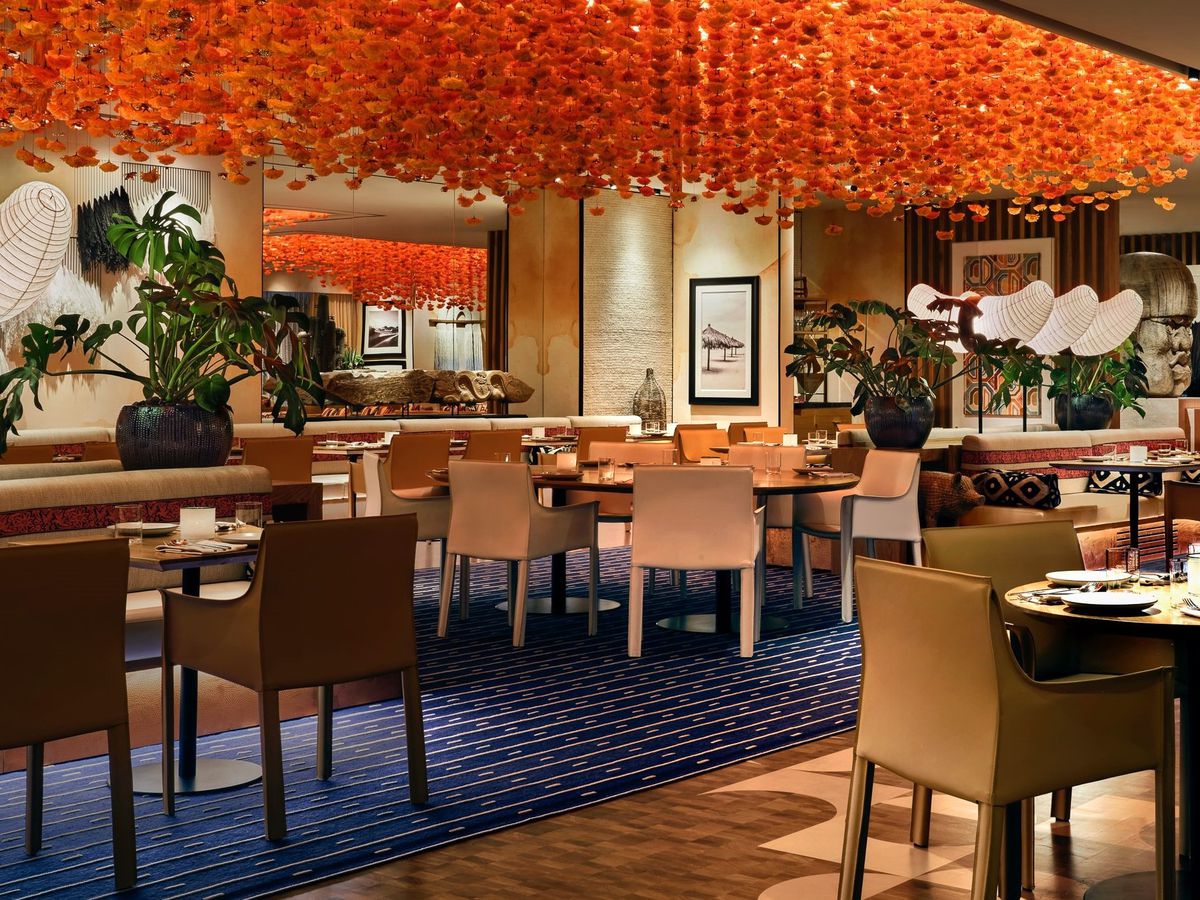 Orange flowers drip from the ceiling in an empty dining room with beige chairs and blue carpet with tropical plants.