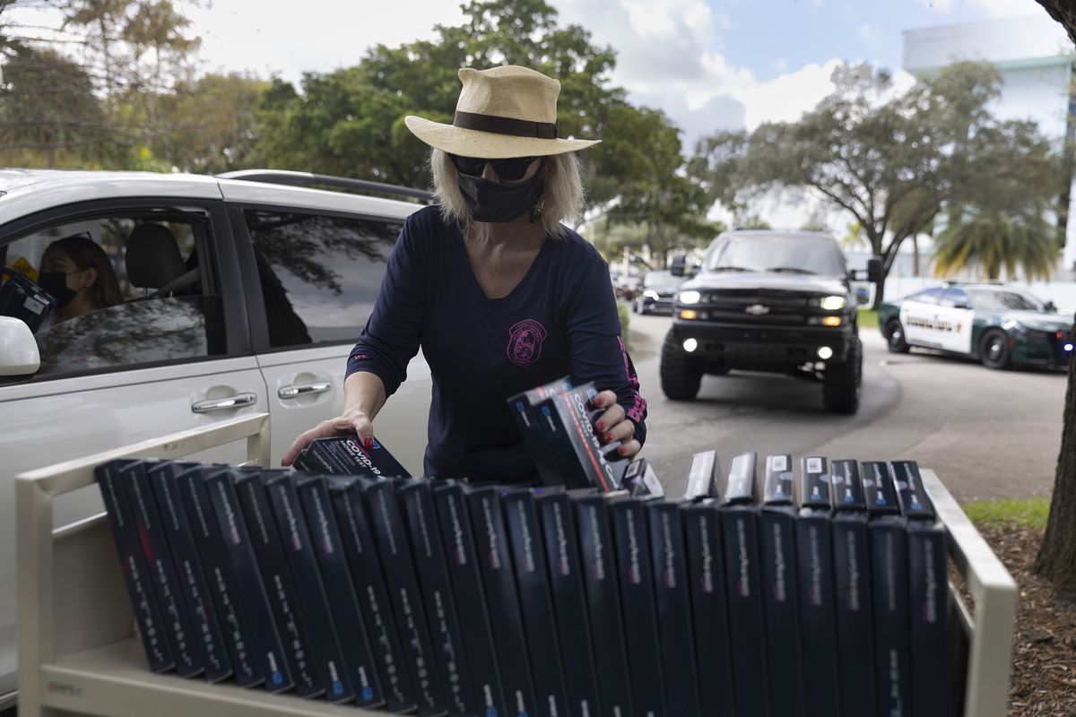 A person hands boxes of rapid tests to people in a car waiting in a drive-through line.