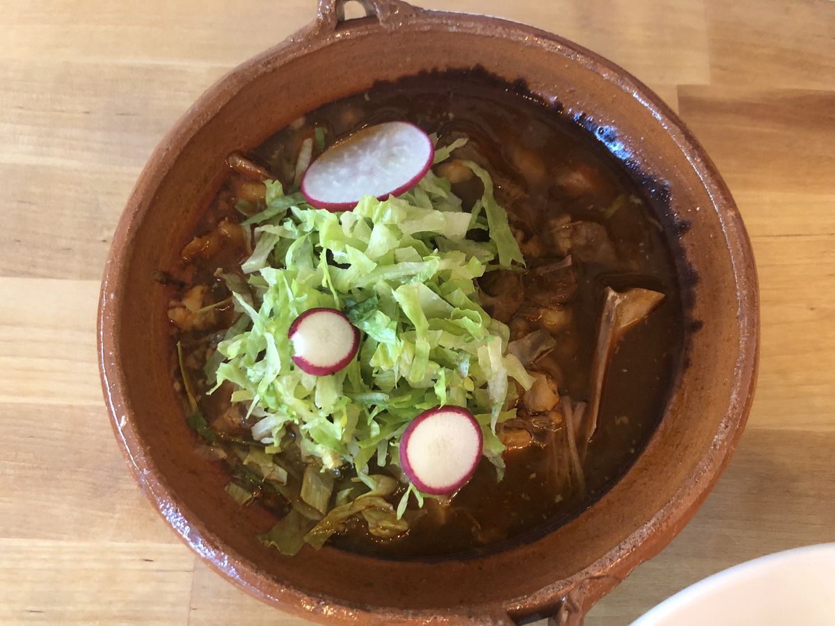 A top-down view of a light-wood table with a dark-orange bowl on it, filled with darkly colored pork pozole rojo topped with bright shredded lettuce and slices of radish.