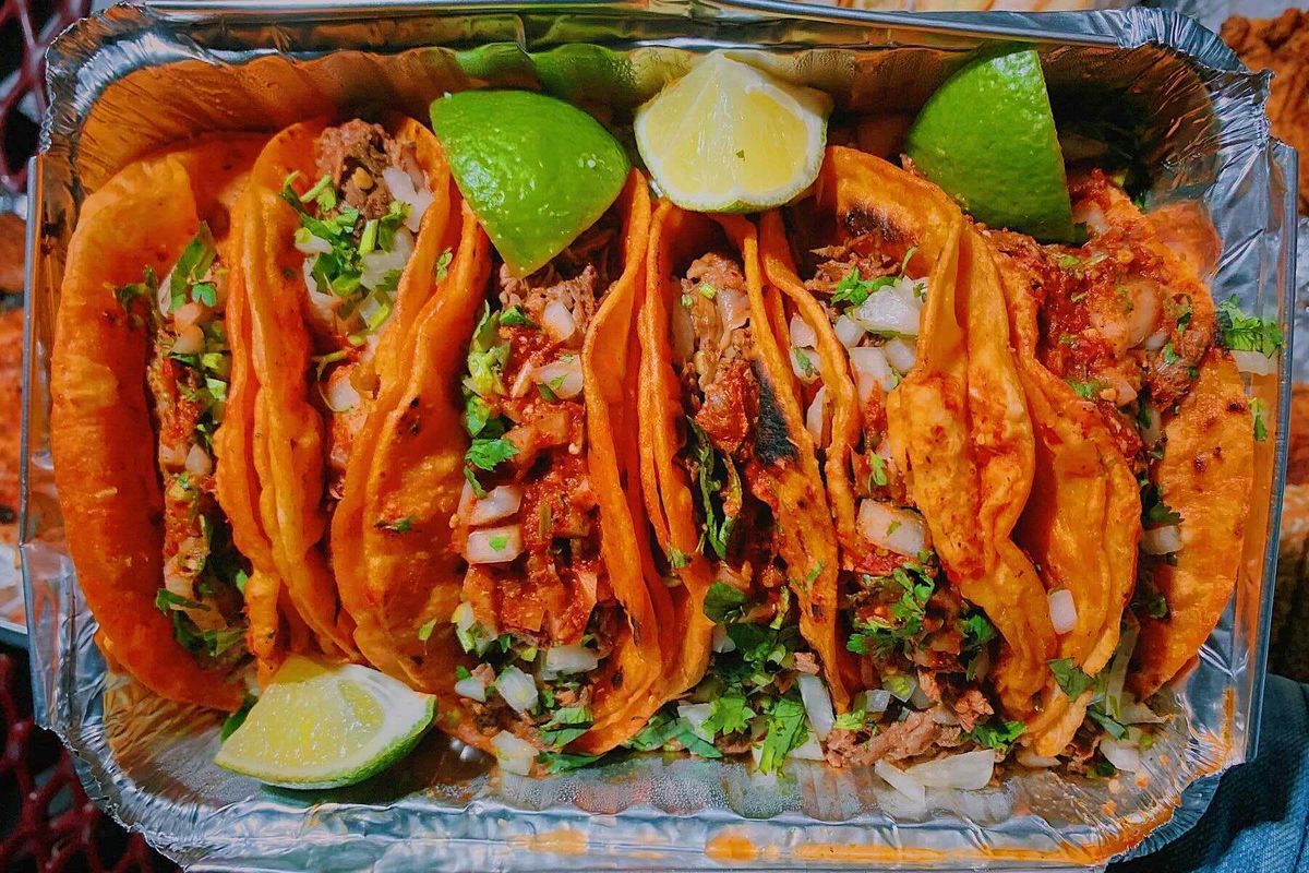 An overhead photograph of six brick-red tacos nestled in a takeout container. Each is double-wrapped in tortillas and stuffed with meat, onion, and cilantro, while wedges of lime dot the exterior.
