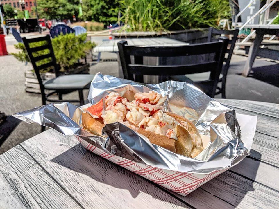 Lobster meat is piled into a hot dog bun, which sits on aluminum foil in a red and white paper holder. It sits on an aged picnic table outdoors, with more tables and chairs, as well as some greenery, visible in the background.