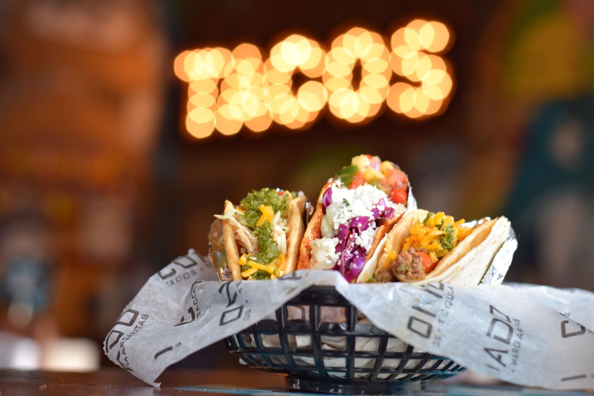 Three tacos in a black plastic basket lined with paper and a lighted up sign reading “Tacos” in the background. 