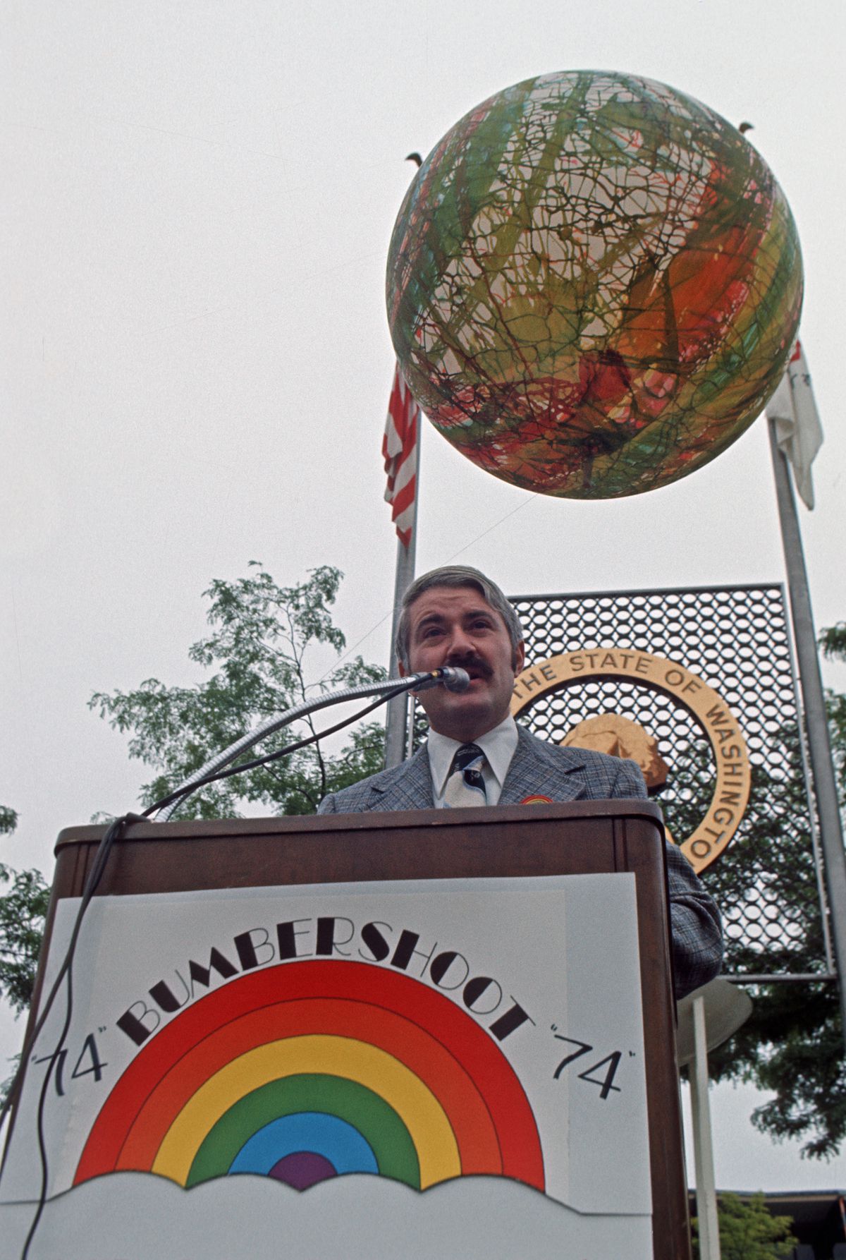 A man with gray hair, a brown mustache, and a gray suit speaks at a podium with a sign attached with a rainbow and the text “74 BUMBERSHOOT 74.”