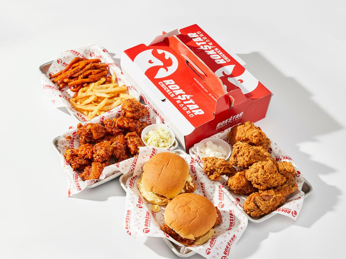 A red and white takeout box and several serving trays filled with fried chicken, two fried chicken sandwiches, and fries.