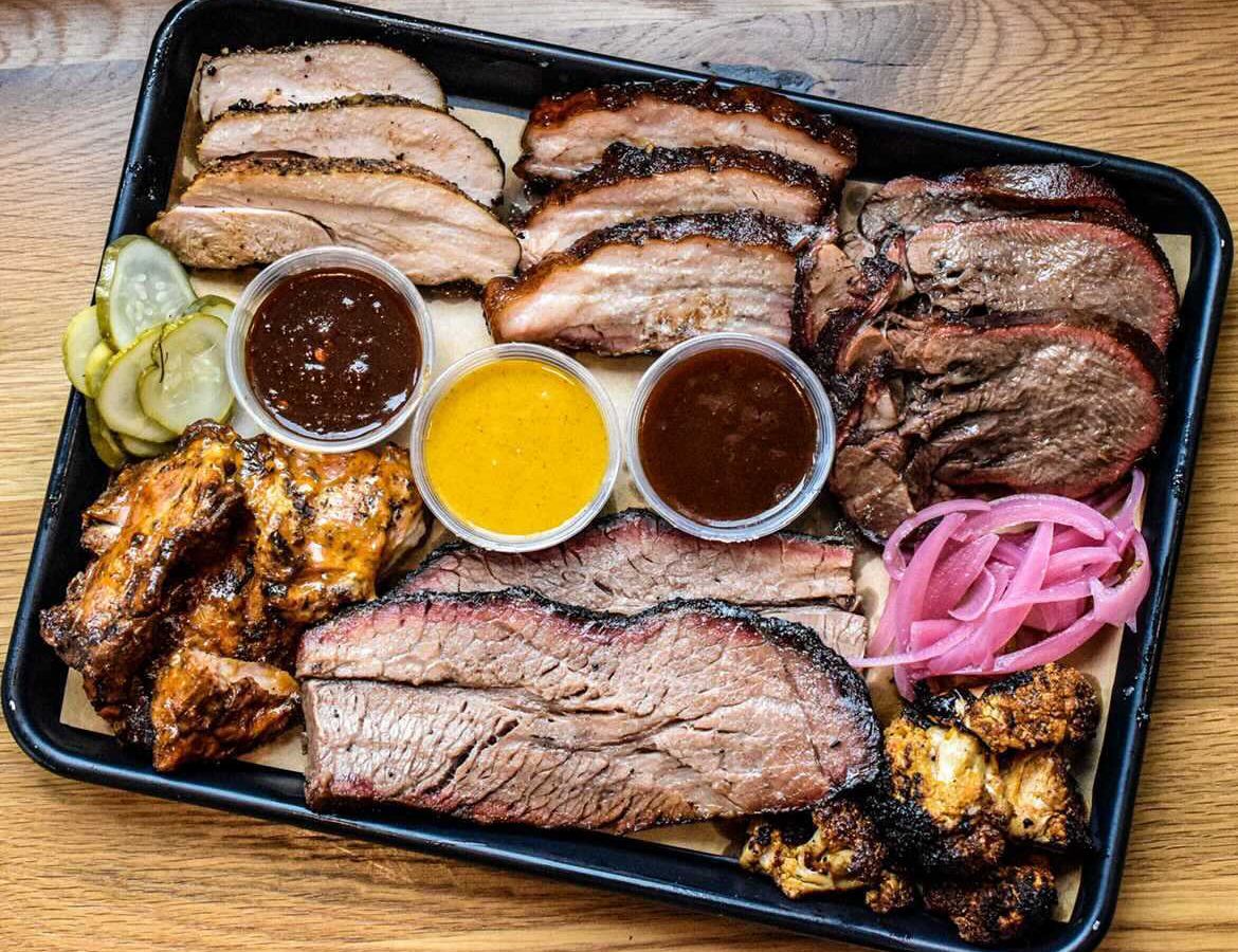 From above, a large tray of slices of various  barbecued meats, separated by variety, along with dipping sauces