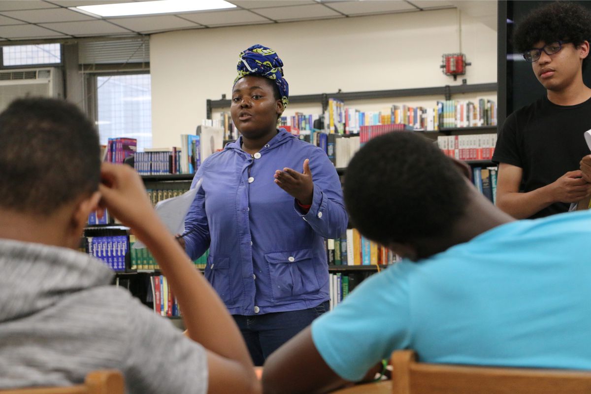 A Black girl in a blue shirt talks to a group of students in a high school. She’s in front of a bookshelf.