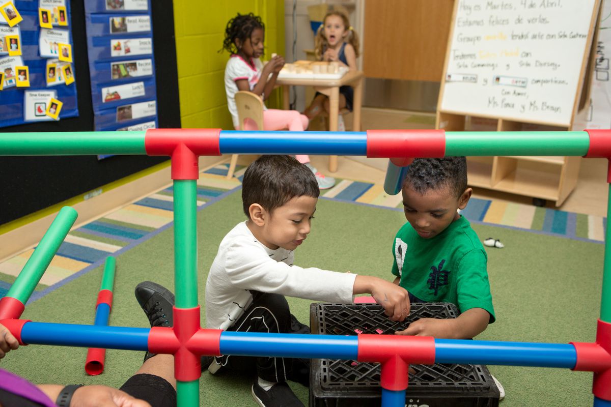 Two preschool boys in the foreground play on a mat in a classroom as two preschool girls sit at a table playing in the background.