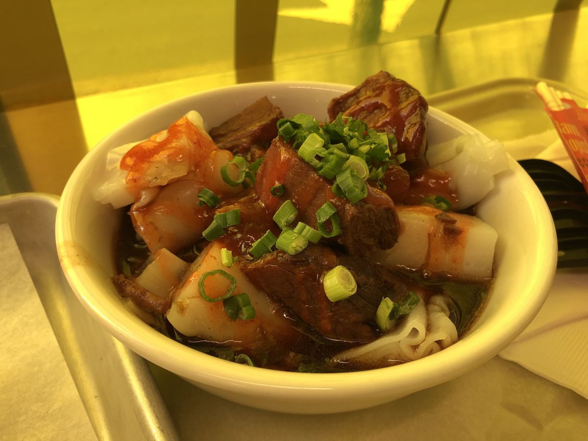 A white bowl filled with white rice noodle rolls, beef brisket, and a dark broth.