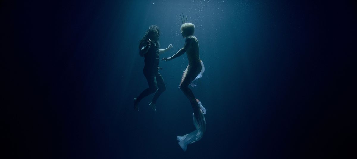 Blackbeard (Taika Waititi) and Stede Bonnet (Rhys Darby, shirtless and in a mermaid tail, carrying a trident) hover together underwater in a deep, featureless blue space in a dream sequence from Our Flag Means Death season 2