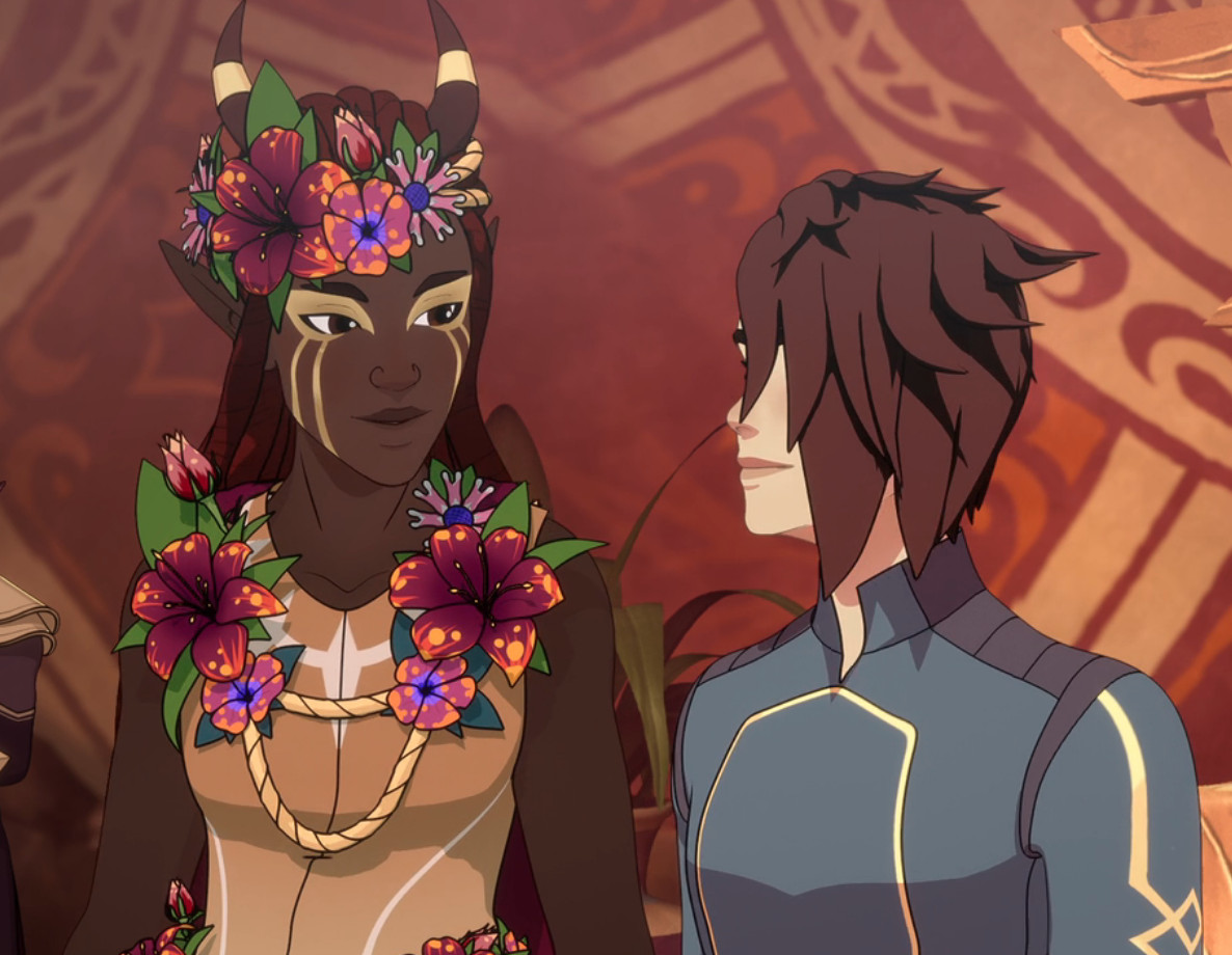 janai, a tall elf woman with dark brown skin and long red hair, stands next to Amaya, a human woman with dark hair in an asymmetrical hair cut and pale skin; both of them are adorned in celebratory flowers and they look warmly at one another