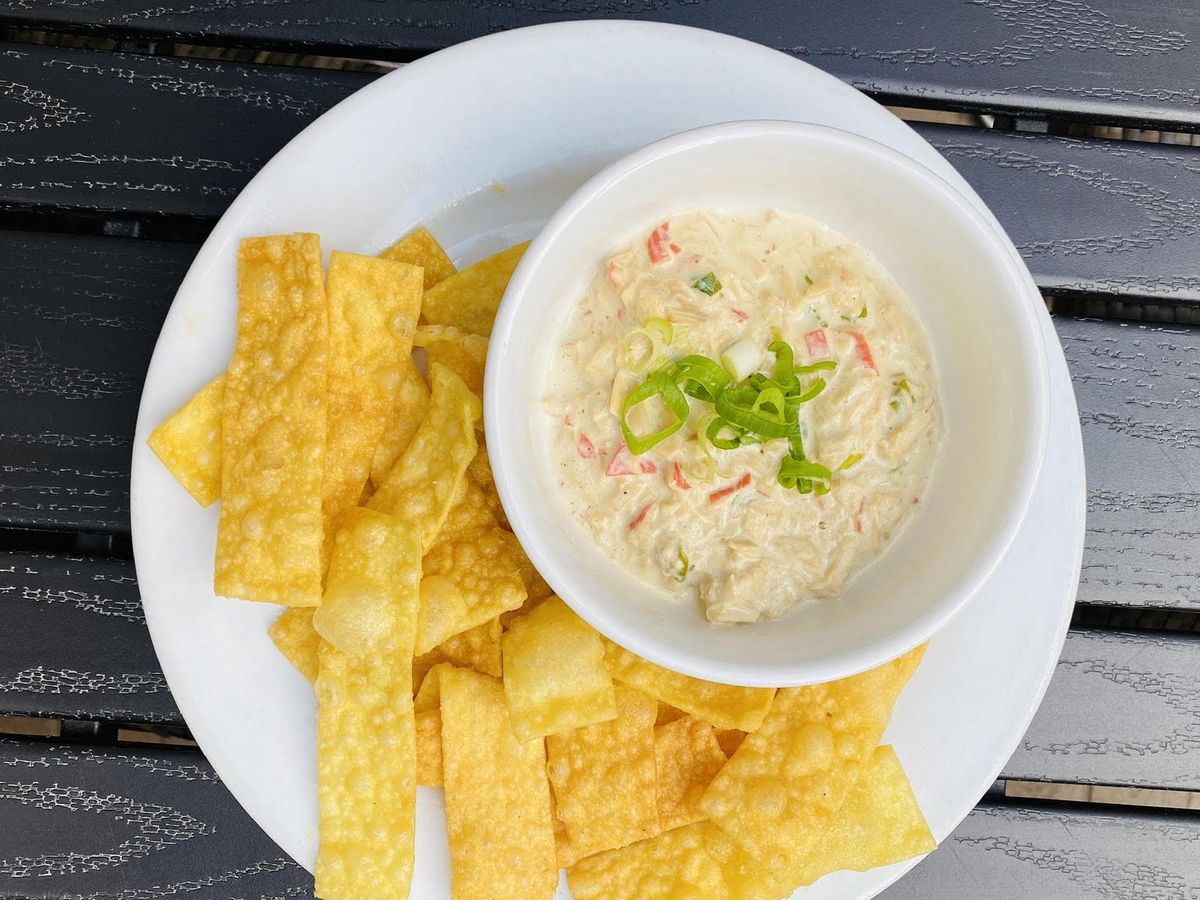 From above, a bowl of crab dip on a plate beside wanton chips, all sitting on a wood-slat outdoor table