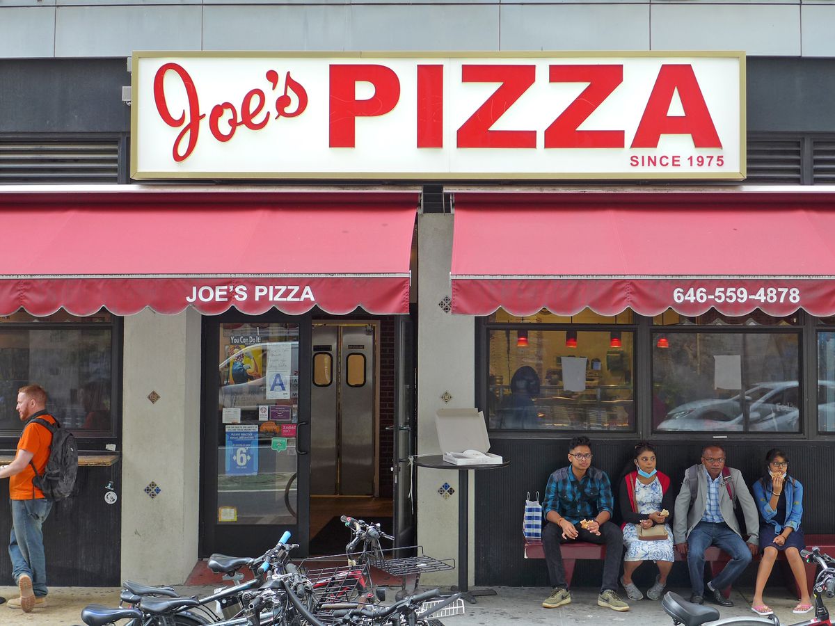 A pizzeria storefront with several people sitting on a bench out front.