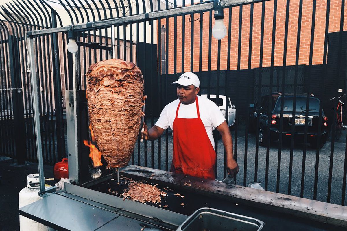 A worker turns a trompo of meat in a red apron.