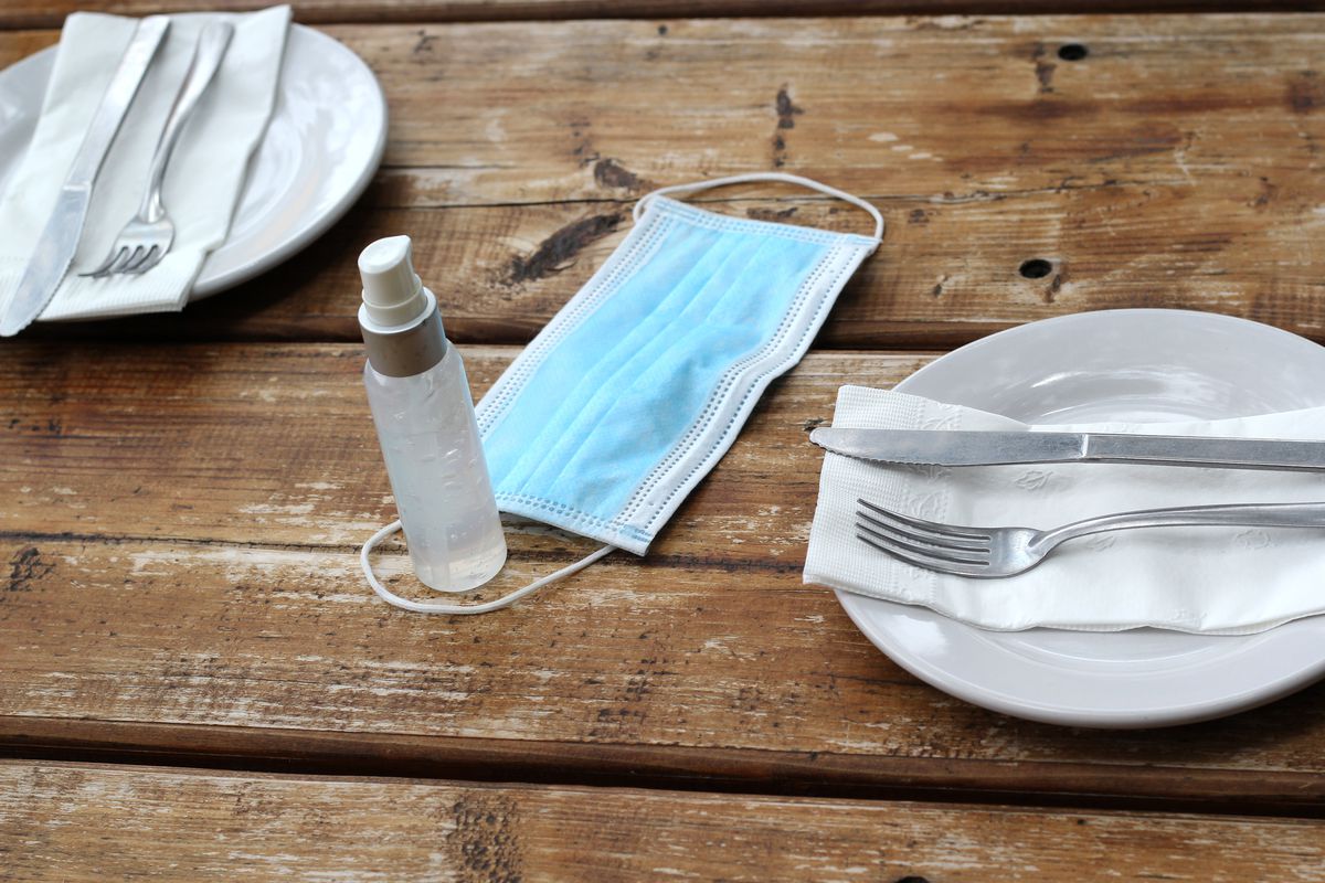 A blue cloth surgical mask sits between two place settings on a wood table next to a small bottle of hand sanitizer.