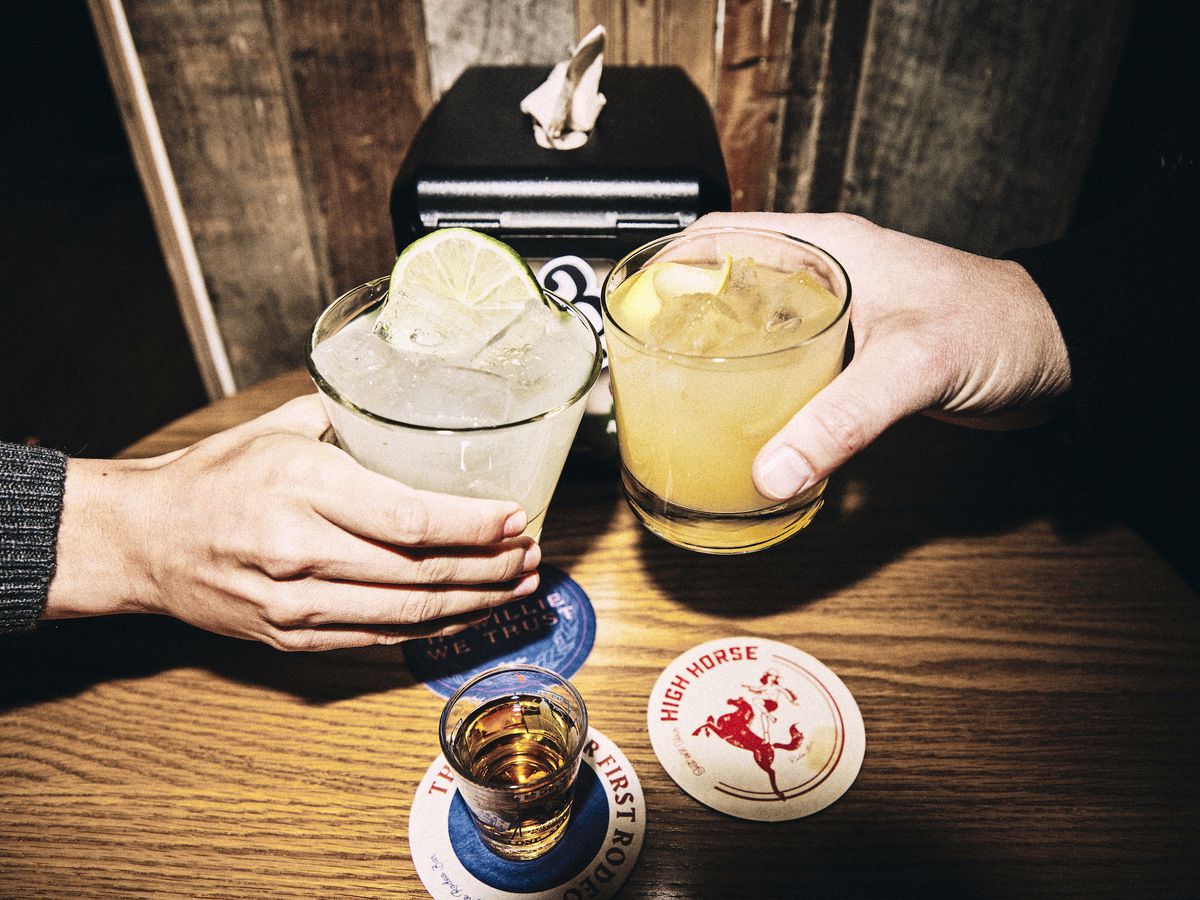 Two hands hold cocktail glasses in a cheers, as a beer and coasters cover the wooden tabletop below.