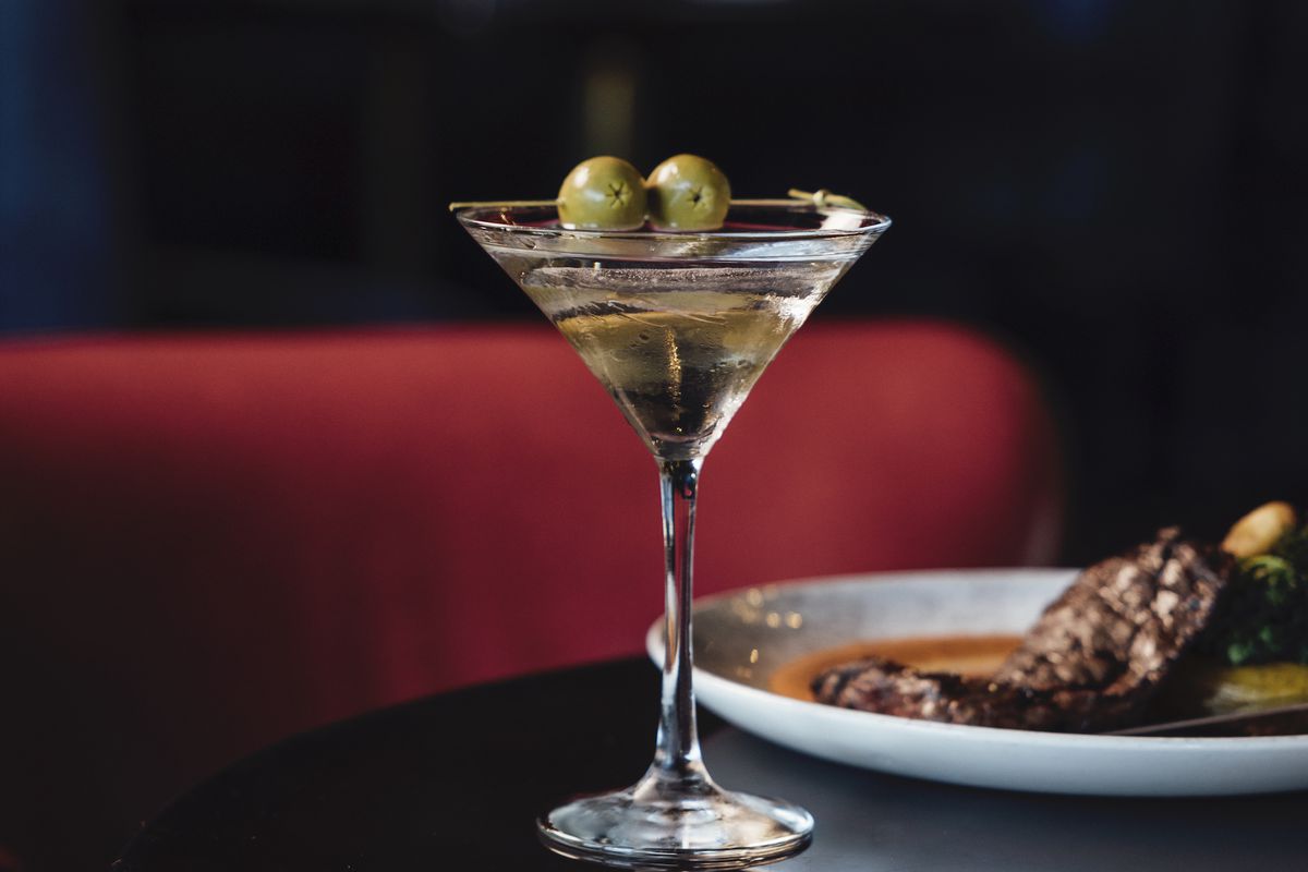 A close up shot of a martini and a seared steak on a white plate, in front of a deep red booth