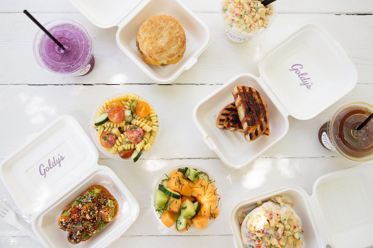 A white table with several food items with some in a white goldy’s takeaway container (avocado toast, purple lemonade, pasta salad, biscuit sandwich, panini, melon salad, cinnamon roll, milkshake, coffee).