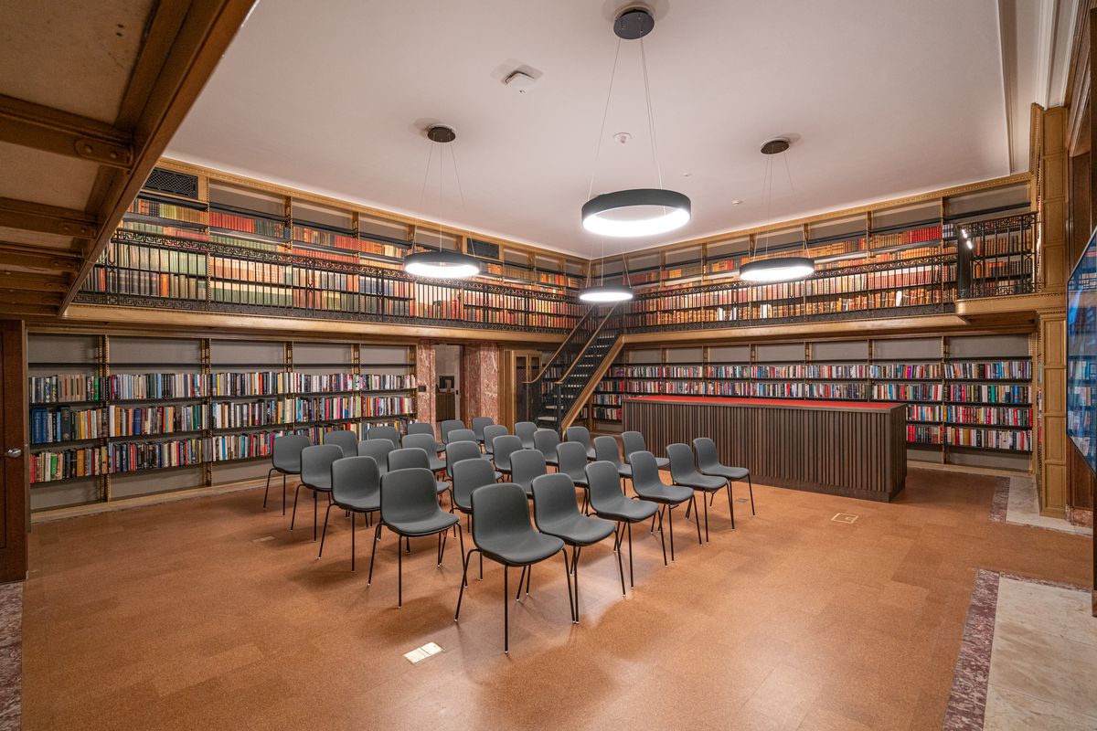A library room with several chairs, wooden floors, and stacks filled with books. 