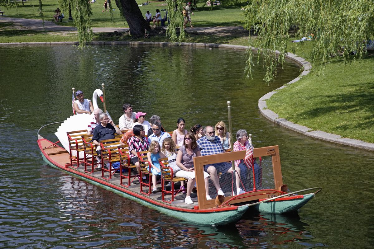 People sitting on chairs on a boat that is traveling through Boston Public Garden.