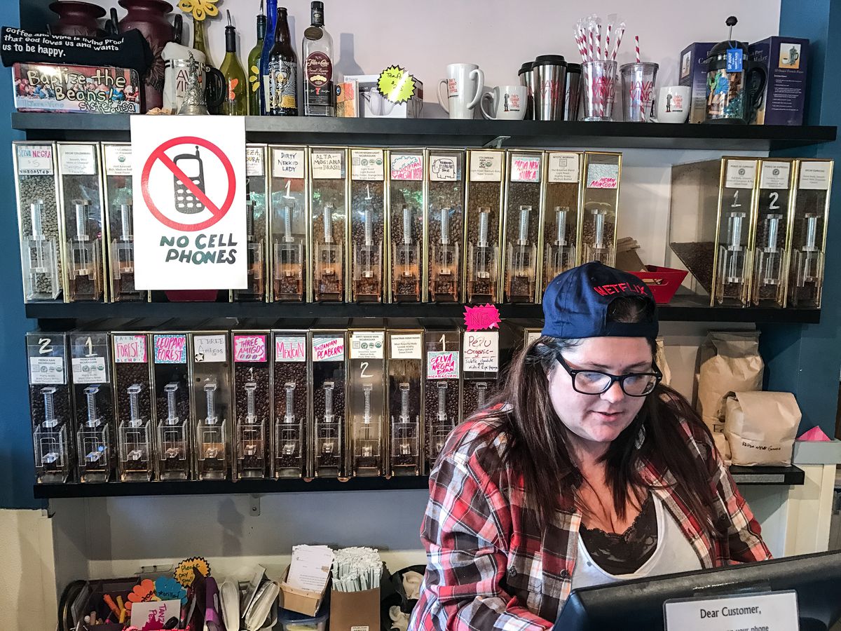 A barista wearing a backwards cap and plaid shirt, and a "no cell phones" sign.
